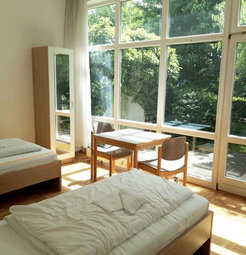 A spacious, sunlit room with two single beds, a wooden wardrobe, a table with two chairs, and large windows revealing lush greenery outside.