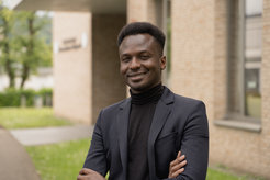Ibrahim Cisse, wearing a dark blue suit and a black high-neck sweater, stands in front of the Max Planck Institute of Immunobiology and Epigenetics in Freiburg, a modern brick building.